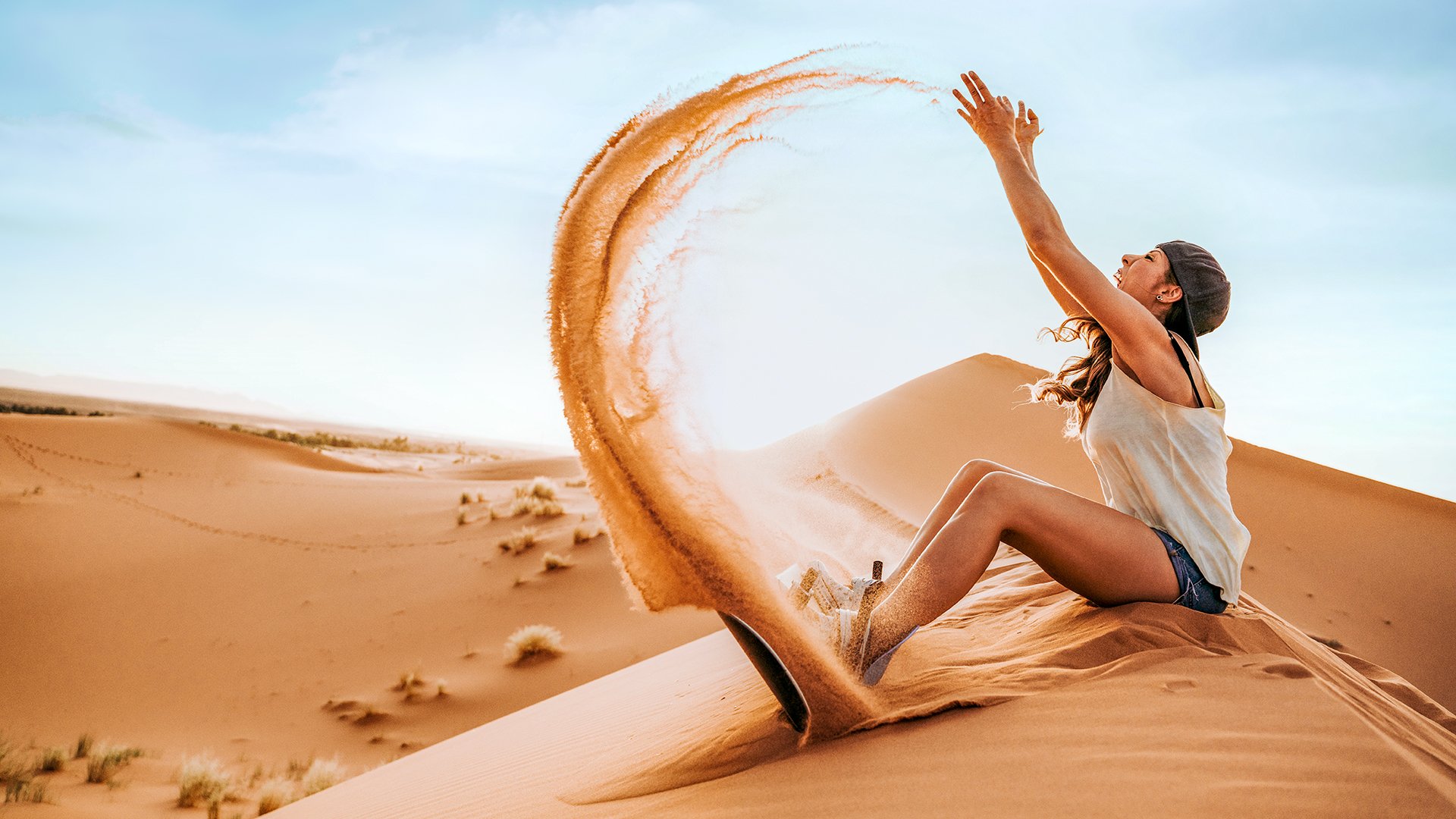 Side view of young female in cap and sunglasses sitting on sand throwing to the air and preparing for sand boarding