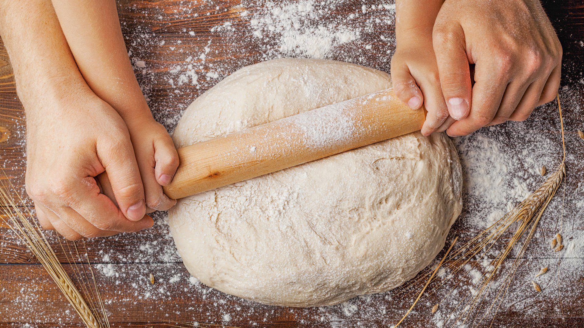 Father and child hands making the dough with flour, rolling pin and wheat ears on rustic wooden table top view. Homemade pastry for bread or pizza. Bakery background.