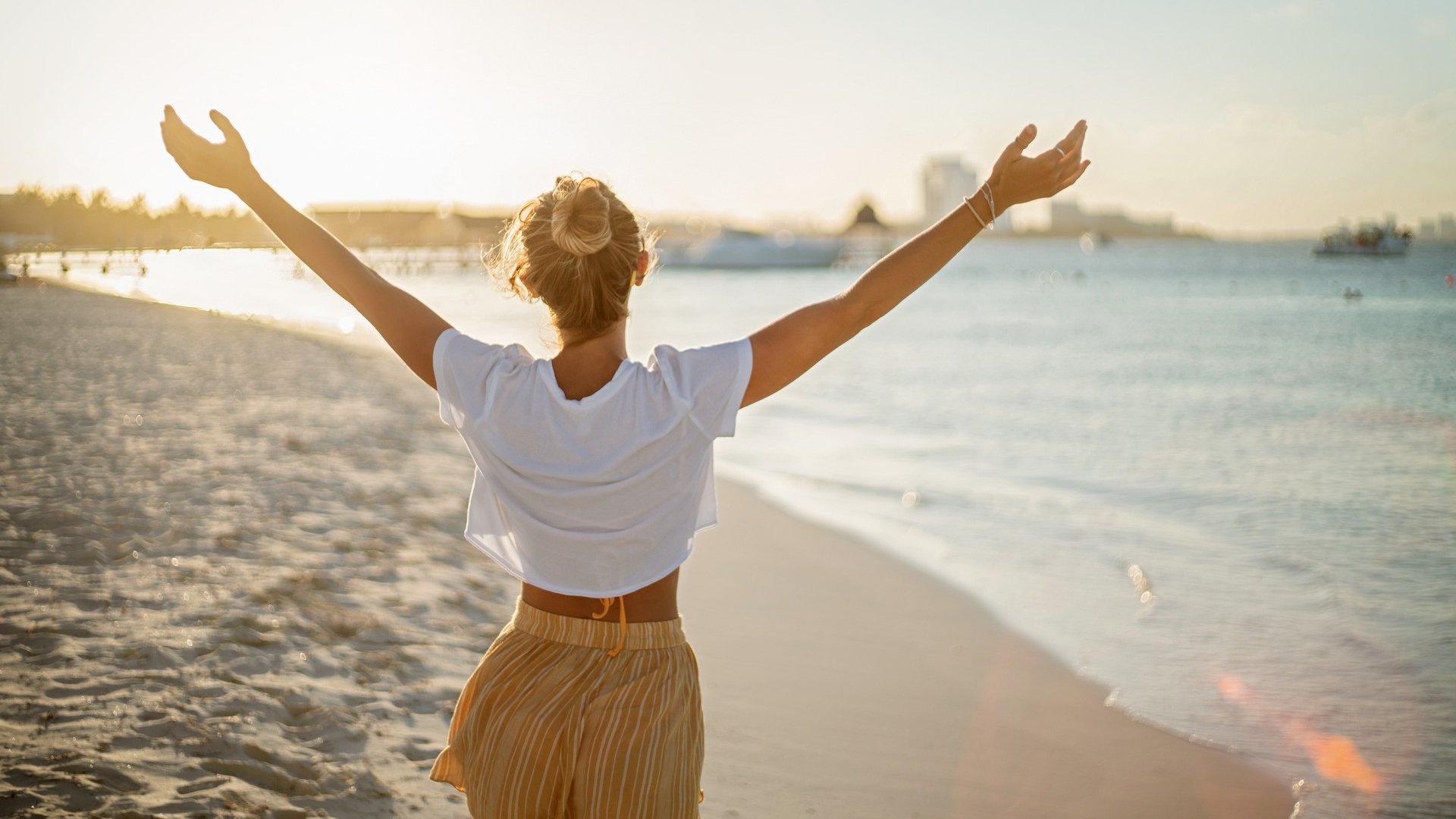 Cheerful young woman embracing nature at sunset; female standing on beach arms outstretched, Cancun, Mexico