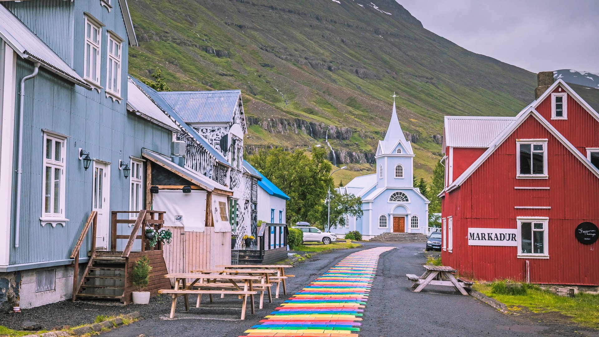 Eine blaue Kirche in Seydisfjördur mit einem bunten Gehweg im Vordergrund und einem großen Berg im Hintergrund..