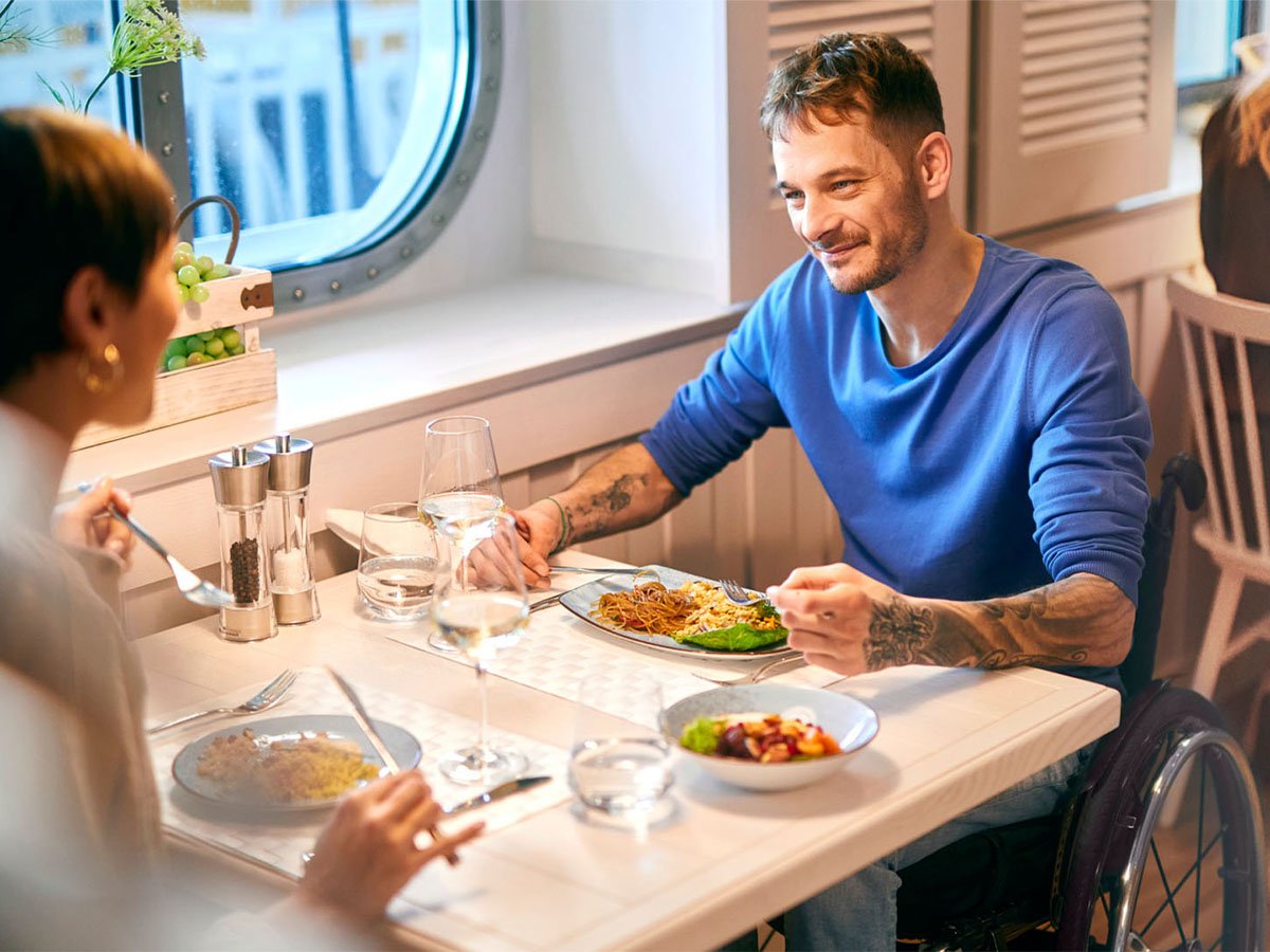 Pärchen sitzt im Restaurant am Tisch und lächelt sich zu. Der Mann sitzt im Rollstuhl, dieser passt genau unter den Tisch im Restaurant.