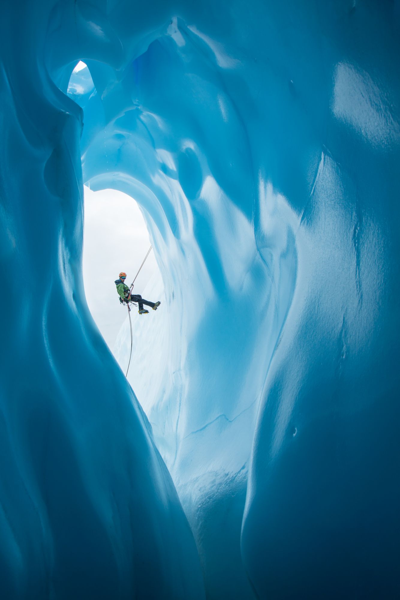Ein Eiskletterer in grüner Jacke und orangefarbenem Helm seilt sich ab an einem großen, runden Eingang einer Eishöhle, Matanuska-Gletscher in Alaska