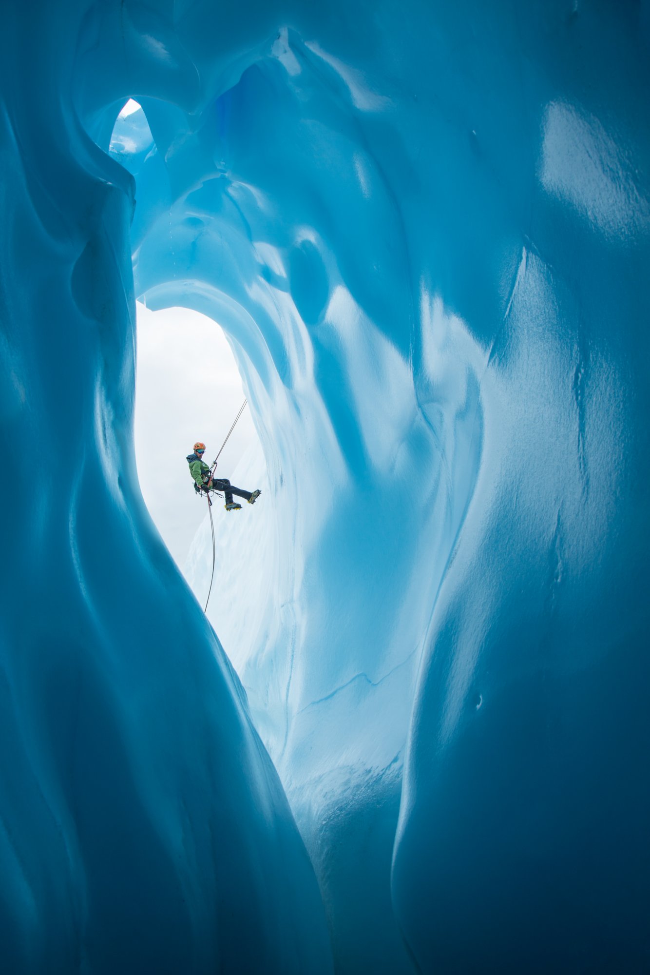 Ein Eiskletterer in grüner Jacke und orangefarbenem Helm seilt sich ab an einem großen, runden Eingang einer Eishöhle, Matanuska-Gletscher in Alaska