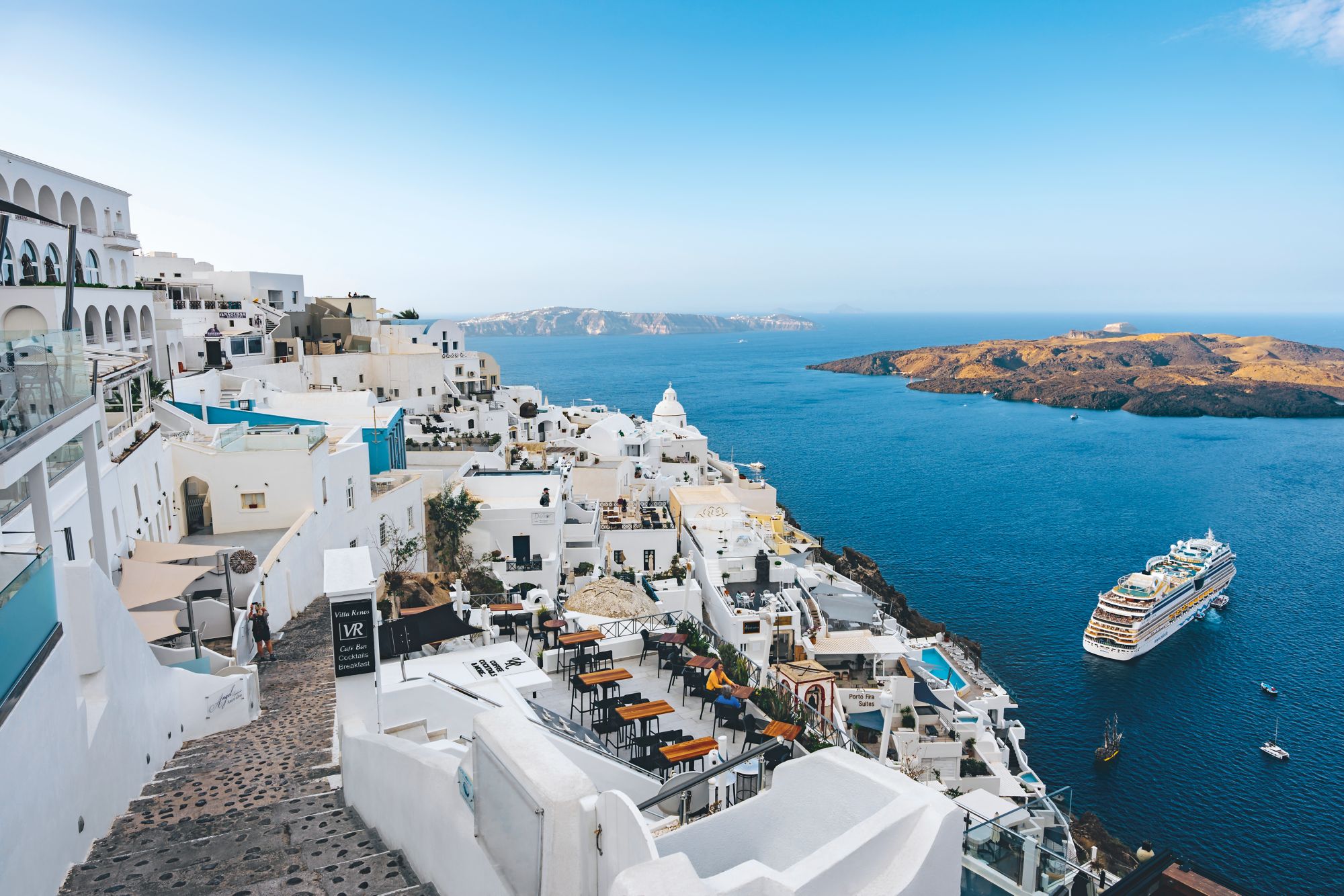 Weiß-blaue Häuser auf Santorini mit Terrassen, Blick auf das Meer und ein Kreuzfahrtschiff vor der Küste.