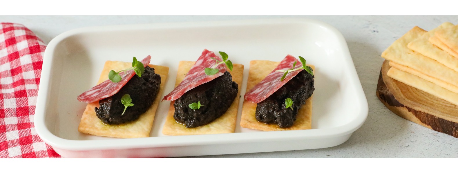 Su un piatto da portata rettangolare sono serviti i biscotti salati con una quenelle di patè di olive e una fetta di salame felino