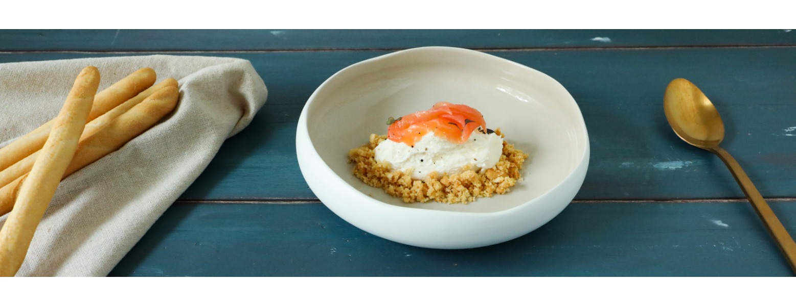 Su un piatto fondo è elegantemente servita la cheesecake scomposta con una base di grissini sbriciolati, una quenelle di crema di formaggi e salmone affumicato