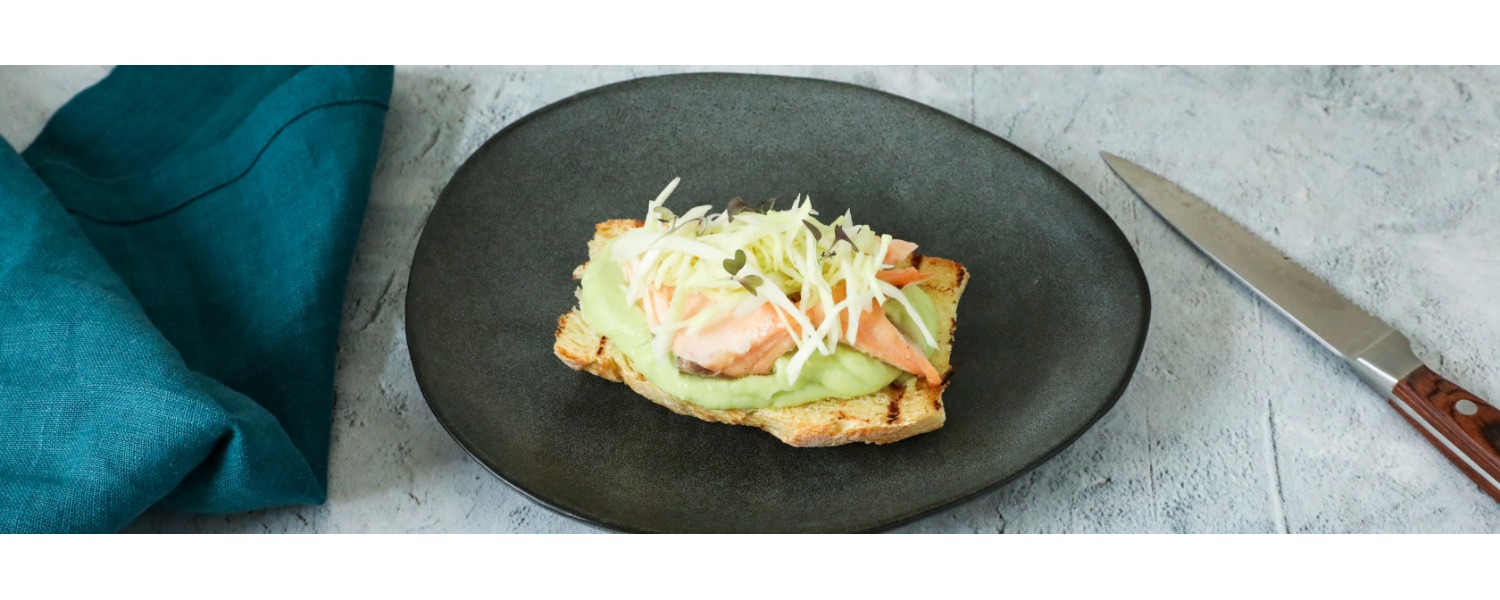 Crostone di pane grigliato con crema di avocado vellutata, salmone scottato e cavolo cappuccio tagliato finemente, servito su un piatto scuro