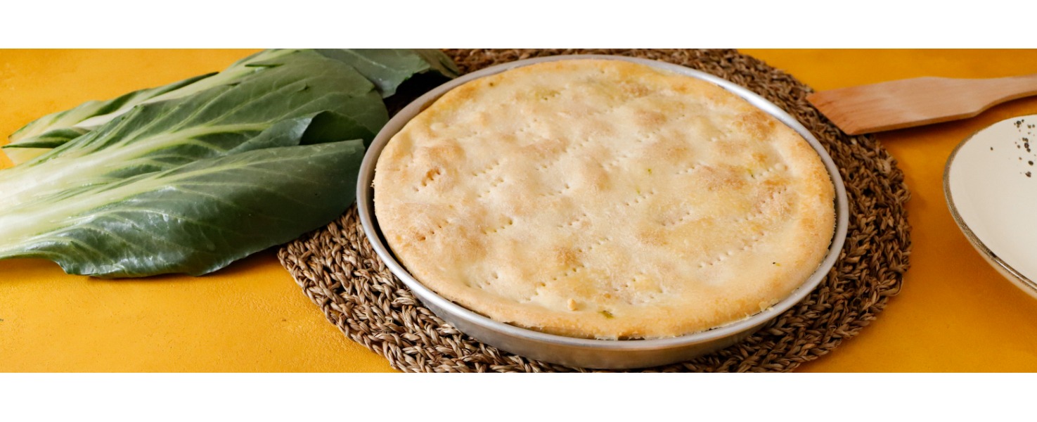 L’erbazzone appena sfornato, nella sua teglia, è poggiato su un sottopiatto di vimini accanto alle biete fresche