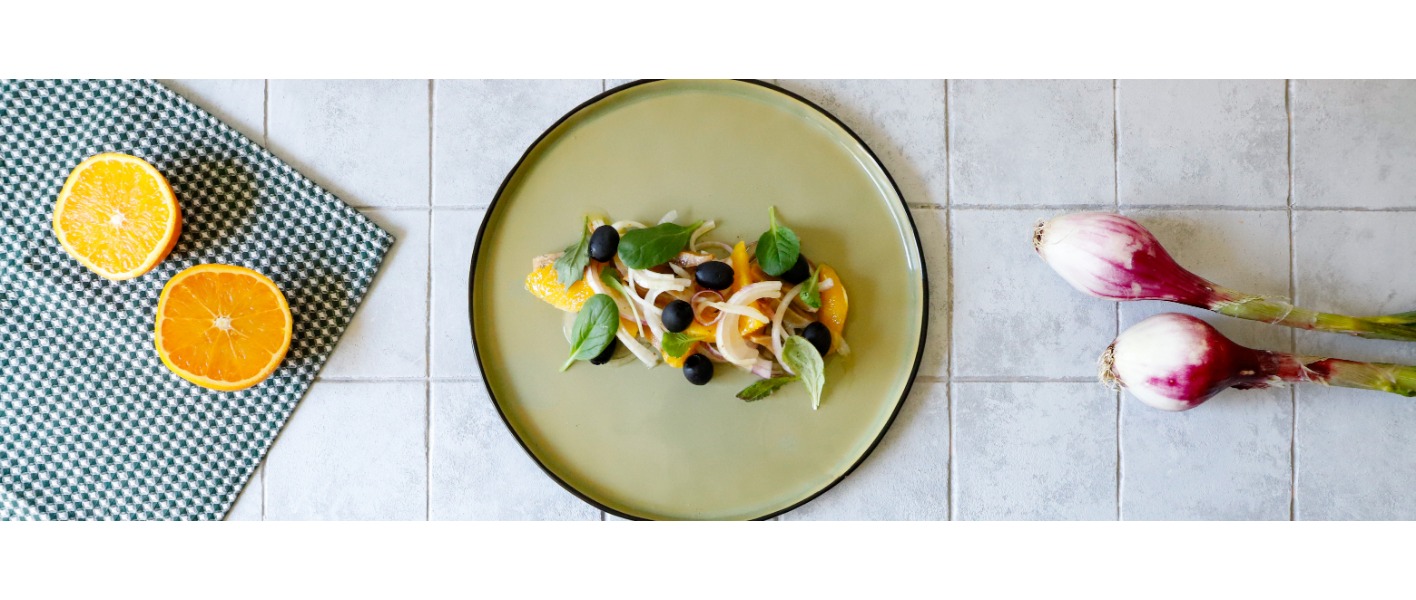 Insalata di aringa è servita su un piatto minimal di colore verde pastello, mettendo in evidenza i colori dell’arancia, la cipolla, le olive e la misticanza