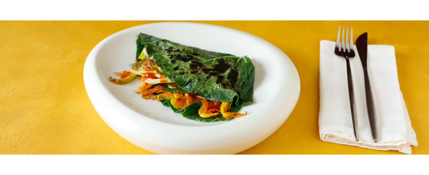 Su un piatto bianco è sistemato un involtino di tenerumi farcito, si intravede un coloratissimo fiore di zucca