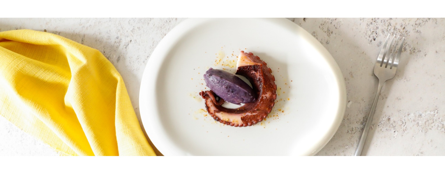 Su un piatto bianco un impiattamento minimal con un tentacolo di polpo e una quenelle di patate viola.