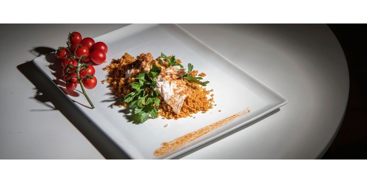 Su un piatto quadrato è servito in maniera elegante il cous cous con zuppa di pesce alla sanvitese e un grappolo di pomodori