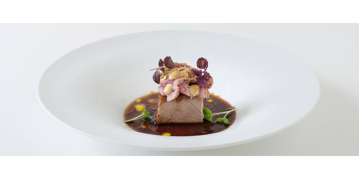 Su un piatto bianco è servito un cubo di ventresca di tonno, fettine di vitello scottato, una crema di capperi e polvere di acciughe
